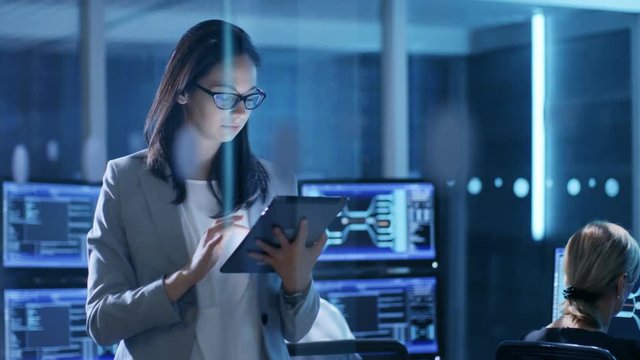 Young Female Government Employee Wearing Glasses Uses Tablet in System Control Center. In the Background Her Coworkers are at Their Workspaces with Displays Showing Data. 