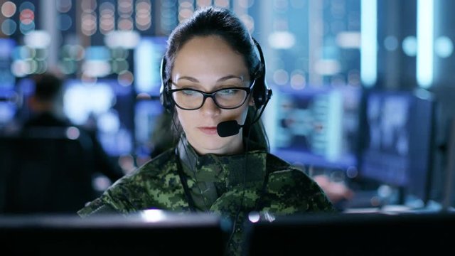 Female Military Technical Support Professional Gives Instructions into Headset. She's in a Monitoring Room with Other Officers and Many Working Displays. Shot on RED EPIC-W 8K Helium Cinema Camera.