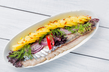 Plate of pieces of herring with fried potato,onion, pepper and herbs on white plate and wooden table