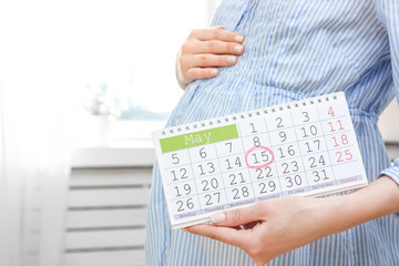 Young pregnant woman holding calendar near belly at home