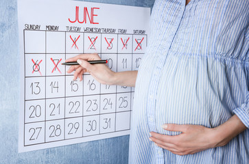 Young pregnant woman counting days with calendar to baby birth on light wall