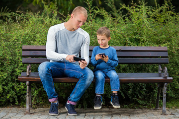 Young father is relaxing on a park bench and playing video games with his little son