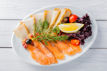 Salmon fillet pieces, sturgeon pieces served with lemon, black olives, herbs, cherry tomato and pomegranate seeds on white plate and wooden table. Top view