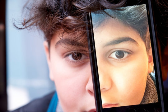 13 year old boys face close up with mobile phone covering one half of his face with a photograph of the other half of his face on it