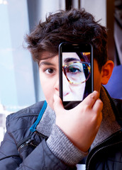 13 year old boys face close up with mobile phone covering one half of his face with a photograph of his mothers half of her face on it