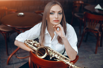 Young attractive girl in white shirt with a saxophone sitting on coffee shop - outdoor in street....