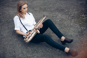 Young attractive girl in white shirt with a saxophone sitting sits on the earth - outdoor. Sexy...