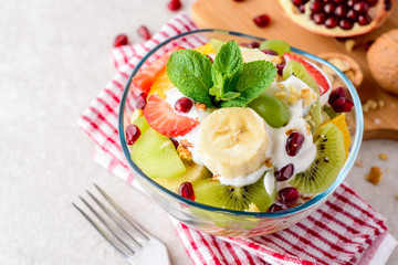 Fresh fruit salad with yogurt and walnuts in glass bowl on stone background.