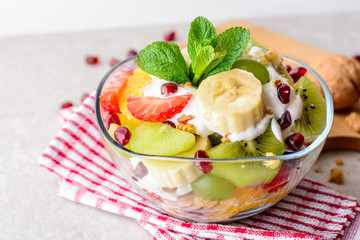 Fresh fruit salad with yogurt and walnuts in glass bowl on stone background.