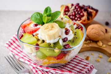 Fresh fruit salad with yogurt and walnuts in glass bowl on stone background.