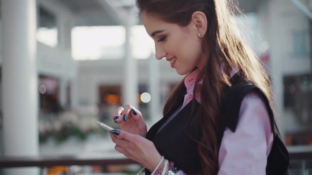 Young smiling woman with beautiful make-up in the pink shirt and black sleeveless jacket is in the shopping center. The girl is concentrated on her smartphone, she browses Internet