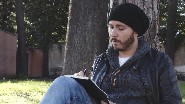 Bearded man sitting under a tree writing in a notebook