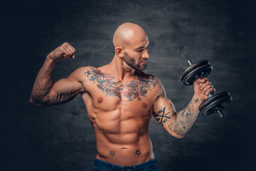 Shaved head sporty male with tattoos on his chest and arms holds dumbbell.