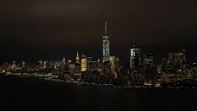 AERIAL HELI SHOT: Skyline view of New York peninsula iconic Downtown Manhattan financial district with famous huge waterfront skyscrapers and giant office buildings lit up with lights shining at night