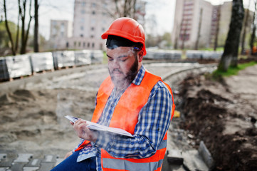 Obraz na płótnie Canvas Brutal beard smoking worker man suit construction worker in safety orange helmet sitting on pavement, break at work, and read working notebook entries.