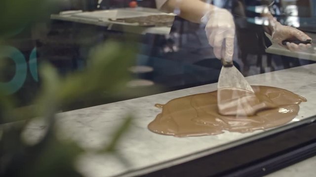 Tracking shot of female confectioner using metal spatula to spread liquid chocolate