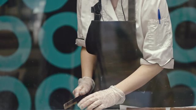 Tilt down shot of woman in apron and protective gloves using knife to cut chocolate fudge into small pieces