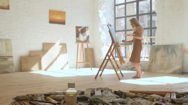 Girl artist paints on canvas on easel and holds palette in art workshop
