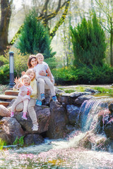A same-sex lesbian family with two children is sitting near a small waterfall in the park. They waved their hands
