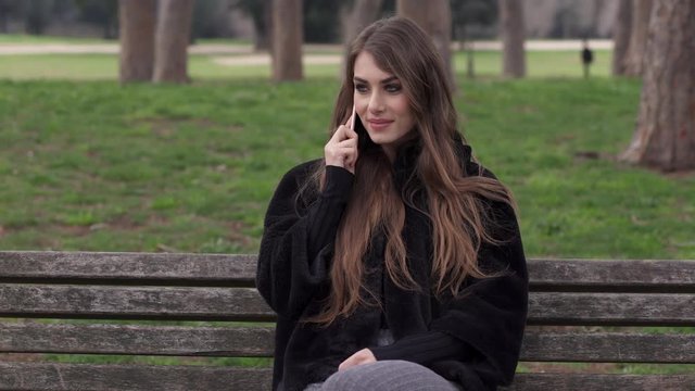 Portrait of cute woman sitting on a park bench, talking on the phone