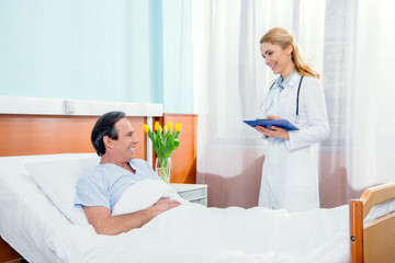 Obraz premium middle aged patient lying on bed and doctor with diagnosis standing near him in hospital