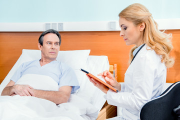 middle aged patient lying on bed, doctor using digital tablet and sitting near him in hospital