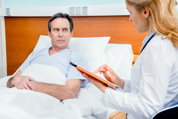 Fototapeta premium middle aged patient lying on bed, doctor using digital tablet and sitting near him in hospital