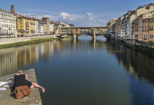 Young blonde female painter leans out of a pylon of the Santa Trinita bridge in the historic center of Florence, Italy, to draw a picture of the famous Ponte Vecchio bridge