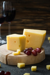 Swedish hard yellow cheese with holes chopped with red grapes on wooden slices and glasses with red wine on dark rustic background