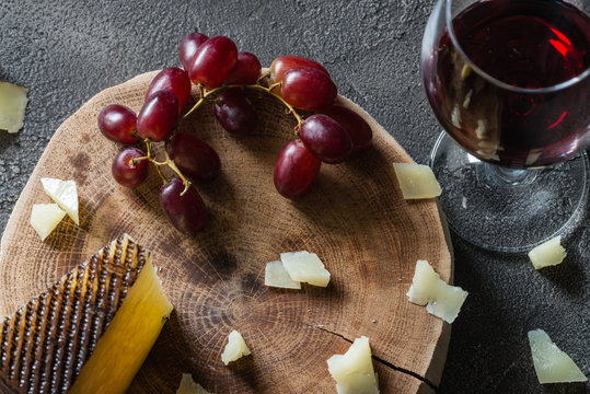 Chopped Spanish hard cheese manchego on wooden cut with red grapes and a glass of red wine on dark rustic background, top view