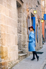 Beautiful young tourist girl in a blue coat and burgundy hat standing in front of the cozy cafe with blue doors and vintage lanterns. Attractive girl with perfect smile.