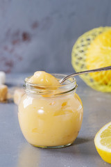 Jar of homemade lemon curd with spoon, whole and sliced lemons, sugar and zest over gray blue stone texture background.