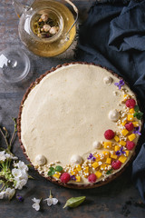 Homemade chocolate tart decorated by mango, raspberries, mint, puffed rice and edible flowers served with glass teapot and blue textile over dark old metal background. Top view. Comfort food concept.