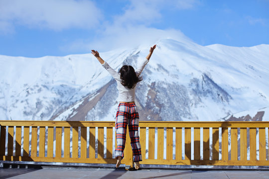 Happy Healthy Brunette Woman In Pajama Back View Stretching On Balcony Overlooking A Mountain Snow Peak, Sunny Morning. Vacation In Resort Hotel. Simple Lifestyle People In Cozy Relaxing Apres Ski