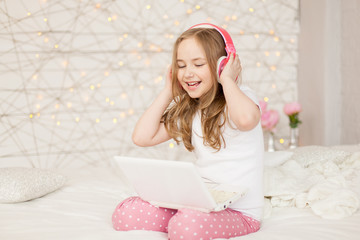 Music and technology. Portrait of young girl in pajamas with white laptop and wireless pink headphones, sing song on background with lights. Pastel colors