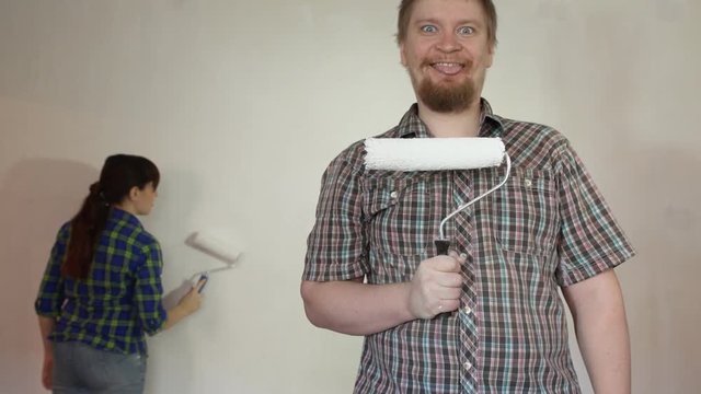 A young red-bearded crazy blond man holds before him a roller with paint, smiles and shows the tongue looking into the camera, against the background of a young woman painting a wall.