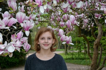 Mädchen steht unter blühenden Magnolienbäumen  ( Magnoliaceae ) und schaut lächelnd nach vorne 