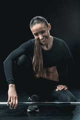 Fototapeta premium young fitness woman sitting at barbell and resting isolated on black.
