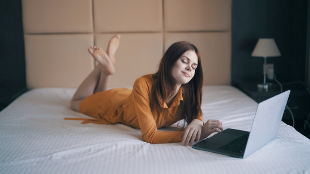 A Woman In A Dress Lies On A Tucked Bed And Looks In Laptop