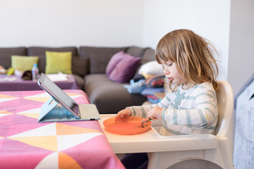 Fototapeta premium Three years old blonde child sitting in white high chair indoor, eating from orange plastic dish, with digital tablet on the table 