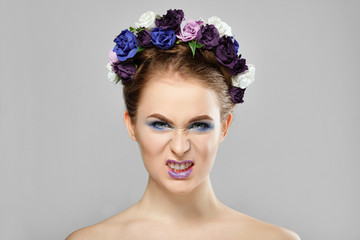Young angry girl with flowers in her hair grins. Perfect creative make up and floral art hairstyle.