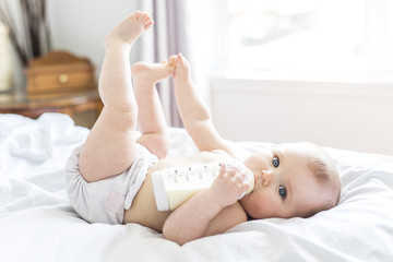 Obraz na płótnie Canvas Pretty baby girl drinks water from bottle lying on bed. Child weared diaper in nursery room.