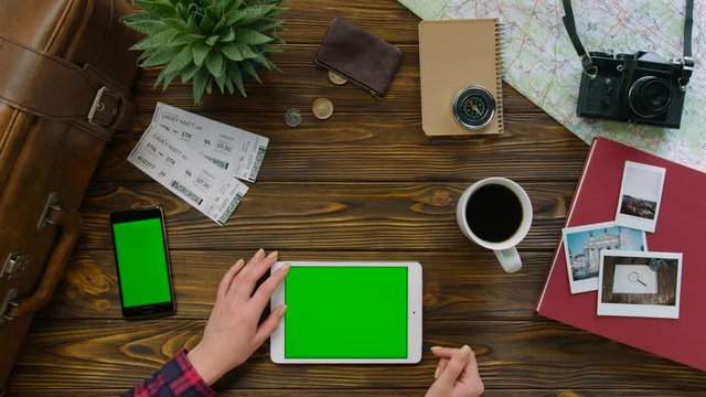 Young woman searching place for the trip on tablet with green screen on wood background with cup of coffee and black smart phone with green screan. Top view. Chroma key