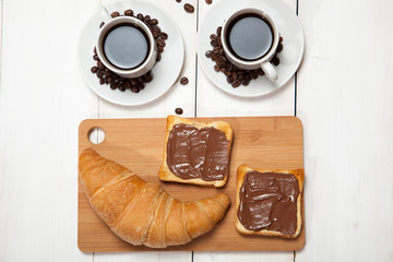 Cups of coffee with a croissant and toast with chocolate on a white wooden table