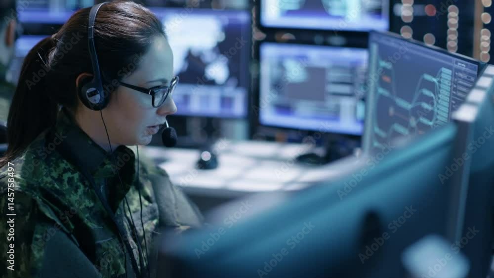 Wall mural Female Military Technical Support Professional Gives Instructions into Headset. She's in a Monitoring Room with Many Working Screens.Shot on RED EPIC-W 8K Helium Cinema Camera.