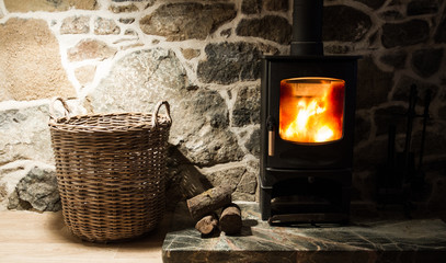 The interior of a cosy, stone cottage with stone walls and a fireplace with logs burning in a wood burner on a fireplace and hearth. - obrazy, fototapety, plakaty