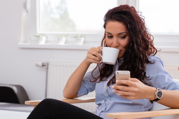Enjoying time at home. Beautiful young smiling woman using mobile phone and drinking coffee while sitting in a big comfortable chair at home