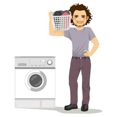 Happy young man holding laundry basket doing chores with washing machine