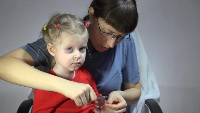 Mother in a blue turtleneck and glasses tonsured nails on the hands daughter in a red T-shirt using pink manicure scissors on a gray background.