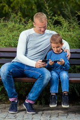 Fototapeta premium Young father and his little son are using mobile phones while relaxing on a park bench